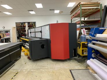 Large industrial printer in a spacious workshop with shelves and rolls of material.