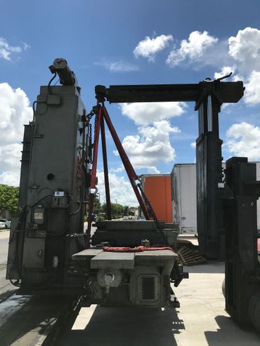 Heavy machinery being lifted outdoors with red straps under a blue sky.