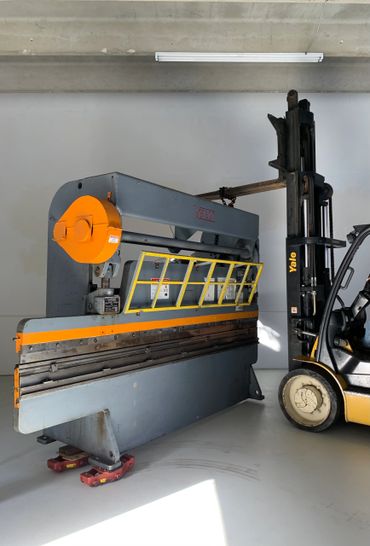 Industrial metal bending machine being moved by a forklift inside a workshop.