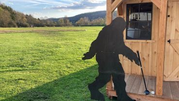 Bigfoot silhouette cutout on a porch with a golf club on grass.