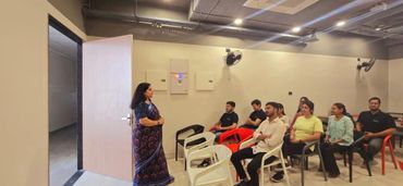 A woman in a saree speaks to a group seated in plastic chairs in a modern room.