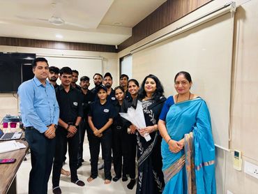 A group of people posing indoors, some in uniform, two women in traditional attire holding flowers.