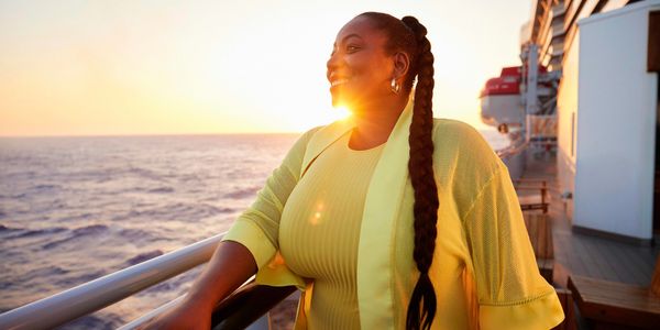 Black woman in yellow dress on the deck of a Virgin Voyages ship looks out over the ocean at sunset