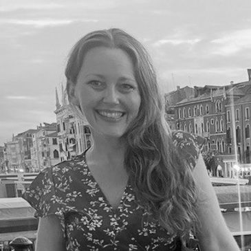 Woman smiling outdoors by a canal with buildings in the background.