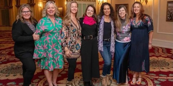 Seven women posing together indoors with smiles and colorful outfits.