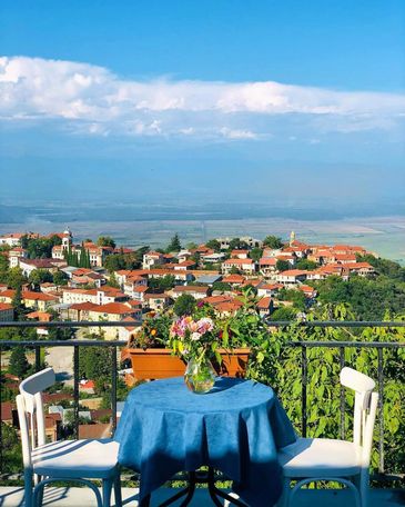 Table with flowers overlooking a scenic hillside village under a blue sky.