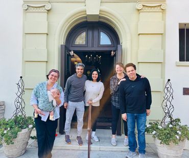 A happy group of five adults and a baby posing outside a building entrance.