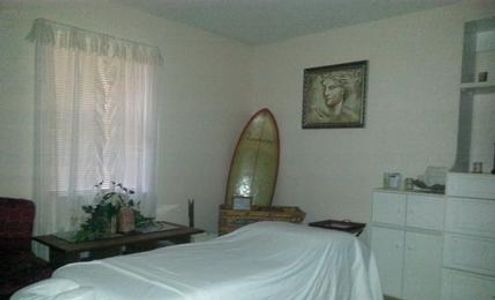 Clean artistic Massage room, white massage table, white cabinets, surf board, chair, window, plant.