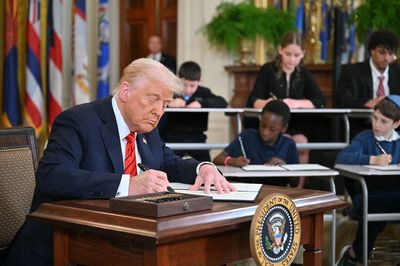Former President Donald Trump signing a document at a desk with children writing behind him.
