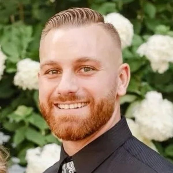 Smiling man with a trimmed beard and patterned tie in front of white flowers.