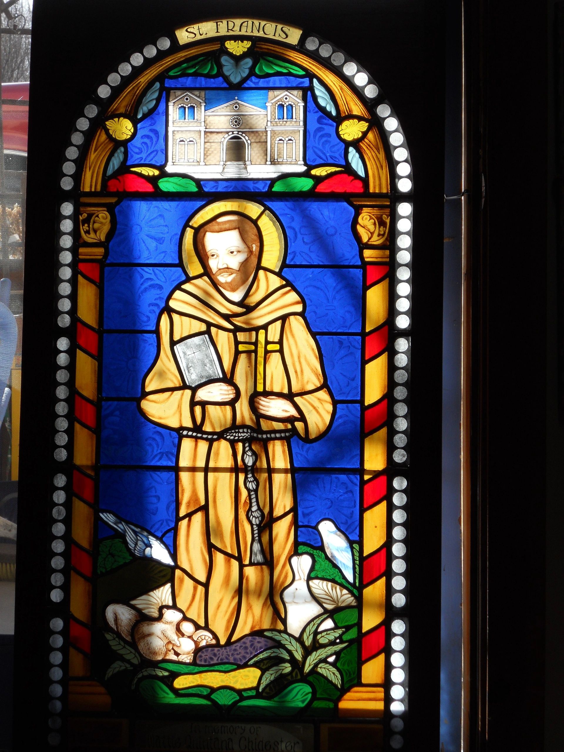 Stained glass of St. Francis of Assisi, painted golden robes against a cobalt blue background with g