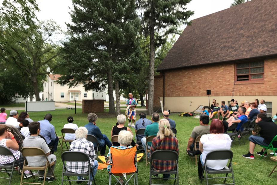 A speaker addressing an outdoor audience seated on chairs.