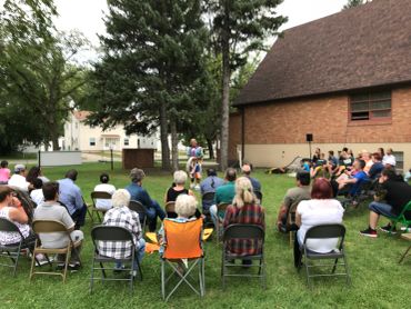 A speaker addressing an outdoor audience seated on chairs.