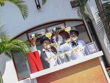 Two men in top hats pouring champagne into a pyramid of glasses outdoors.