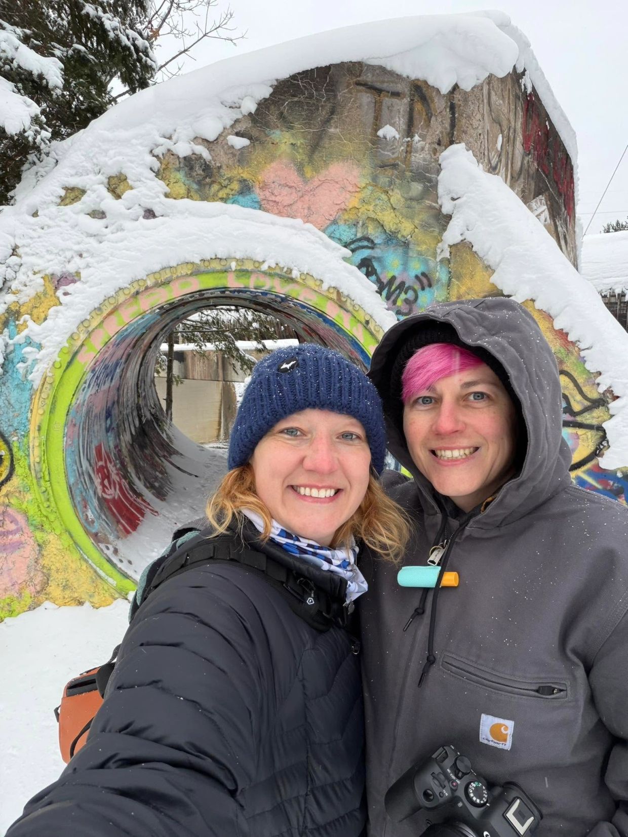 Two people smiling in front of a graffiti-covered snow tunnel.
