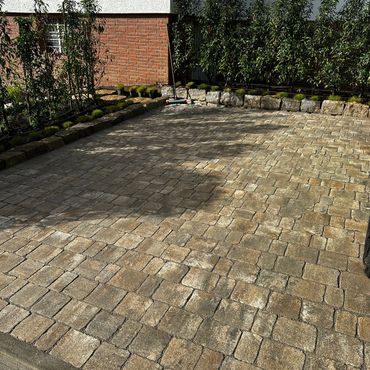 A clean cobblestone driveway with bordering bushes and a broom resting against a wall.