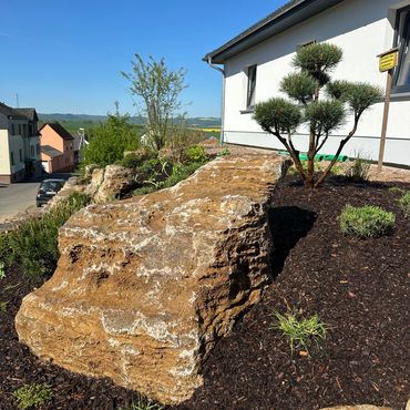 Rock garden with plants beside a white house under a clear blue sky.
