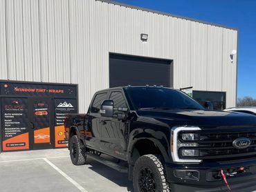 Black Ford Super Duty truck parked outside a window tint and wrap shop on a sunny day.