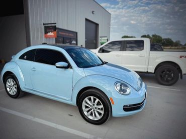 Light blue Volkswagen Beetle parked beside a white truck in an outdoor lot.