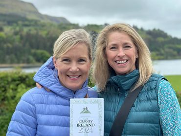 Two smiling women outdoors holding a "Journey Ireland 2024" brochure.