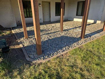 Backyard patio area with gravel, wooden posts, and stepping stones under a covered porch.