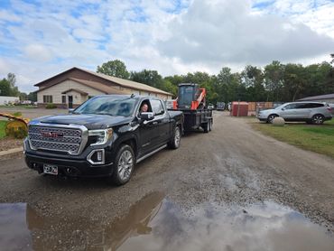 Black GMC truck towing a trailer with construction equipment on a cloudy day.