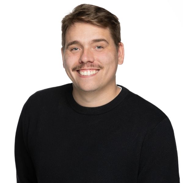 Smiling man with a mustache wearing a black sweater against a white background.