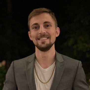 Smiling man in a gray blazer with a gold chain at night.