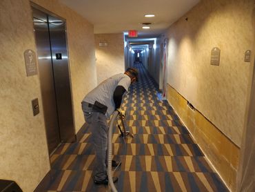 A man cleans a patterned carpet in a hotel hallway near an elevator.