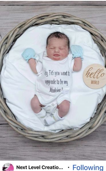 Newborn baby in a basket with a message on the onesie asking to be a Madrina.
