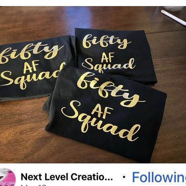 Black shirts with gold lettering saying 'fifty AF Squad' folded on a wooden table.