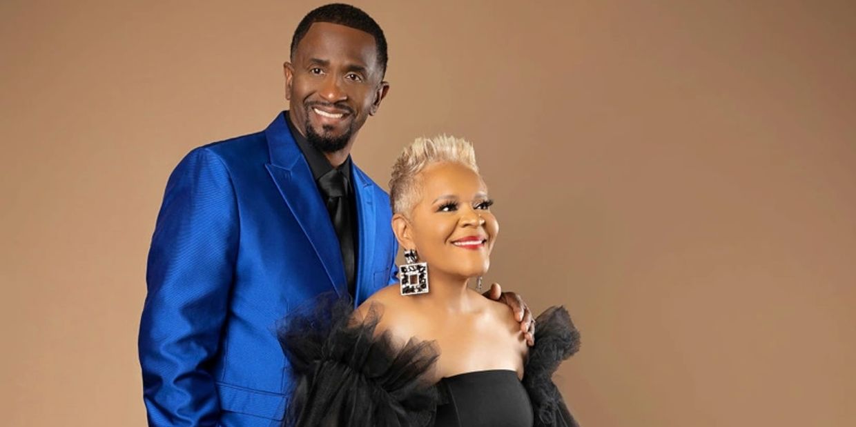 A smiling couple dressed elegantly, the man in a blue suit and the woman in a black dress with statement earrings.