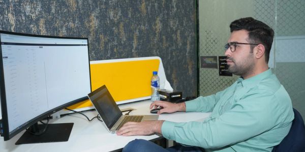Man working on a laptop with a large curved monitor in an office.