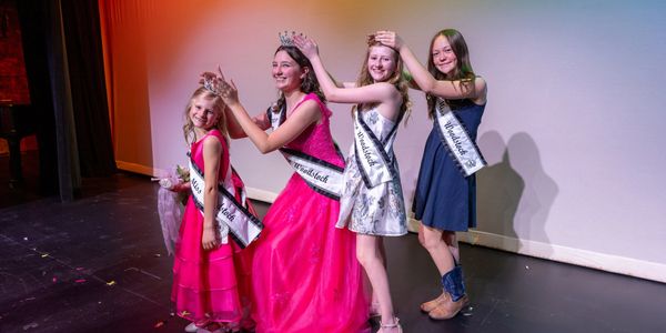 Four girls in pageant dresses and sashes, crowning each other on stage.