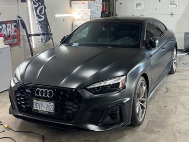 Matte black Audi sedan parked inside a garage with a white wall and boxes in the background.