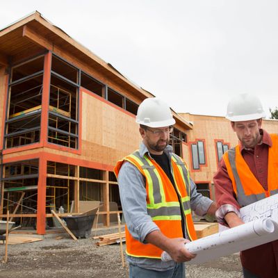 Photo of two construction men in hardhats looking at paper plans with a partially constructed buildi