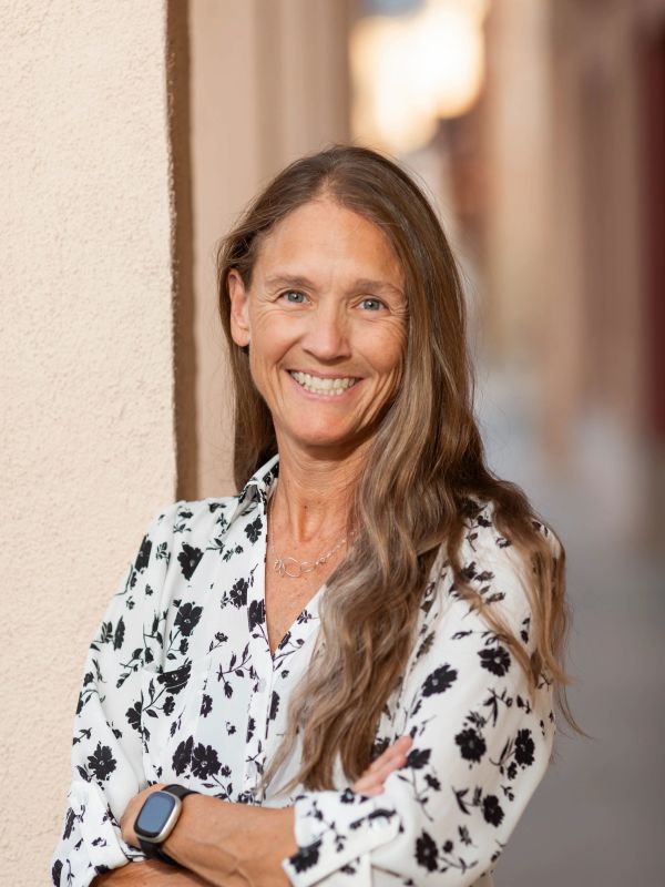 Smiling woman with long hair and floral shirt leaning against wall.
