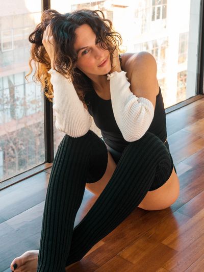 Woman with curly hair wearing black bodysuit and long socks, sitting by window.