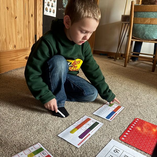 A young boy sitting on the floor solving math worksheets with a marker.