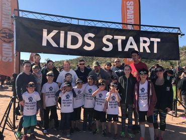 Group of kids and adults under a 'KIDS START' banner at a race event.