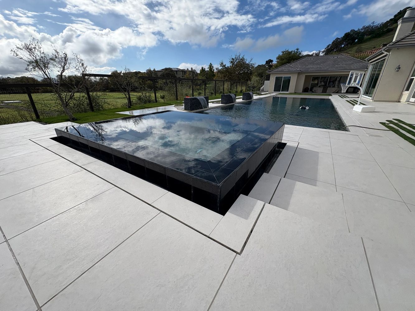 Modern backyard with a pool, spa, and basketball hoop under a partly cloudy sky.