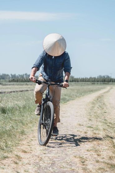 Man wearing a conical hat rides a bicycle on a dirt path in a rural area.