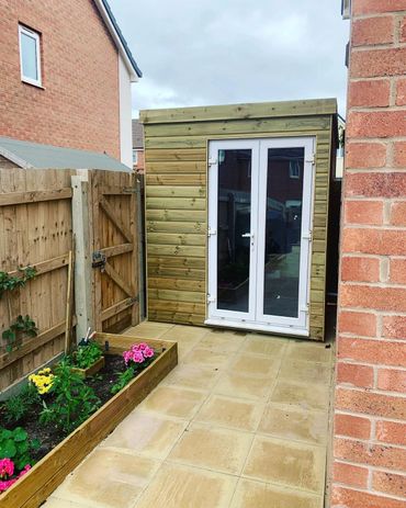 A small wooden garden shed with glass double doors next to a flower bed and a wooden fence.