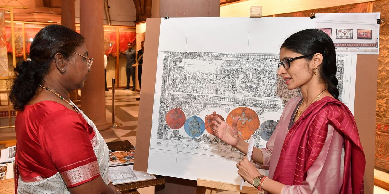 Two women discuss a detailed artwork on an easel in a traditional setting.