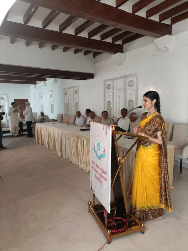 A woman in a yellow saree speaks at a podium in a formal meeting room with seated men.
