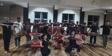 Group of dancers posing in traditional attire with musicians holding drums indoors.
