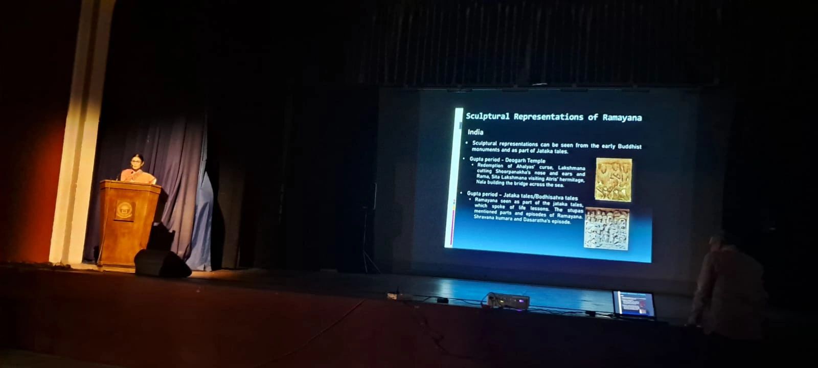 A speaker presents a lecture on Ramayana's sculptural representations in a dimly lit auditorium.