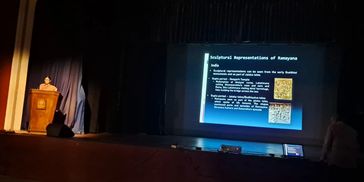 A speaker presents a lecture on Ramayana's sculptural representations in a dimly lit auditorium.