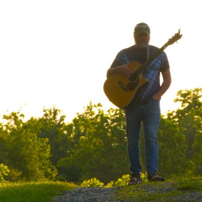 Man walking with a guitar on a path at sunset.