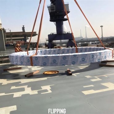 A large ring wrapped in protective material is lifted by straps at an industrial site.